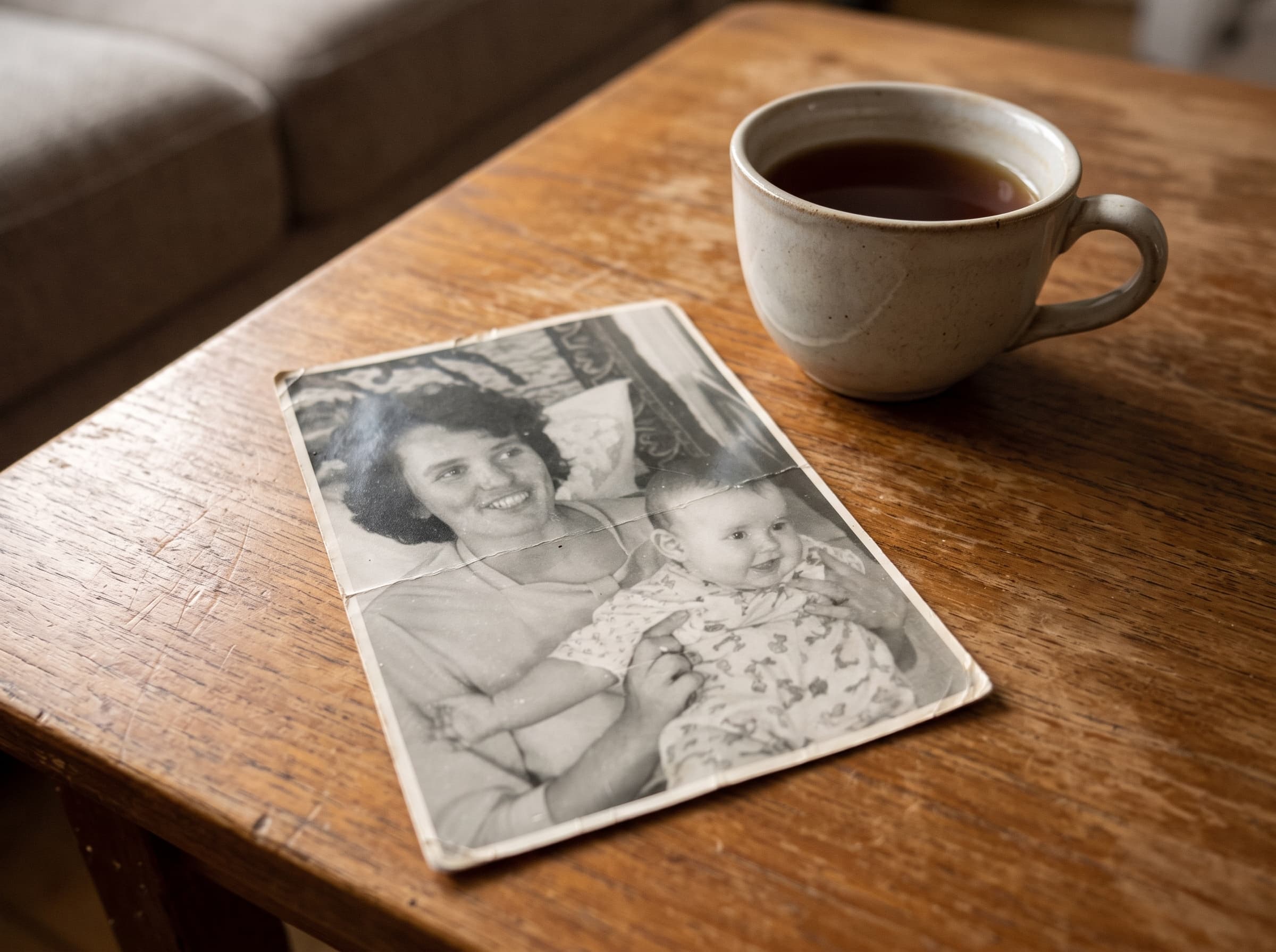 The original black-and-white snapshot — a mother holding her baby — creased and lying on a wooden table beside a cup of tea
