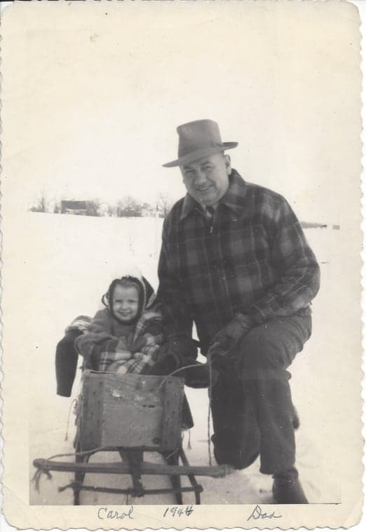 The original photo from 1944: a grandfather kneeling in the snow beside a little girl on a wooden sled